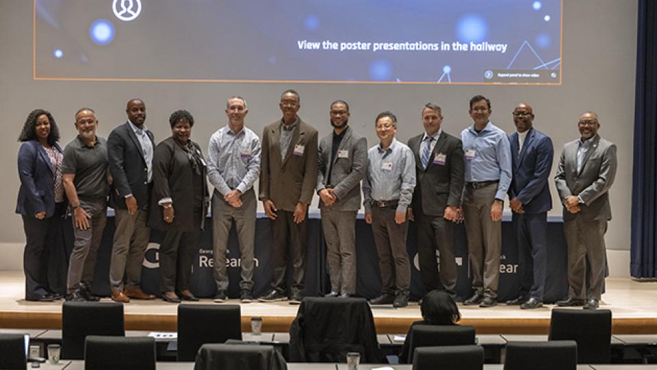 Group of professionals standing on a stage in front of a large presentation screen displaying agenda and seed grant booklet information at a conference.