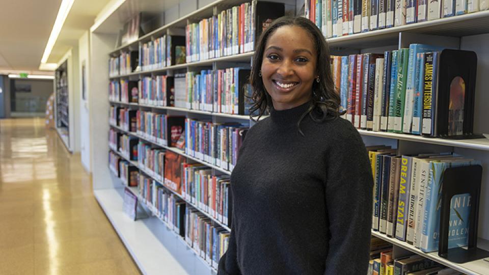 Person standing in a library aisle next to shelves filled with colorful books, wearing a dark sweater and jeans.