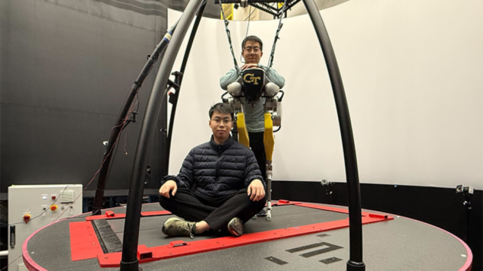 Two individuals on a large circular motion platform in a research lab, with one person seated cross-legged on the platform and another suspended in a harness wearing a Georgia Tech exoskeleton system.