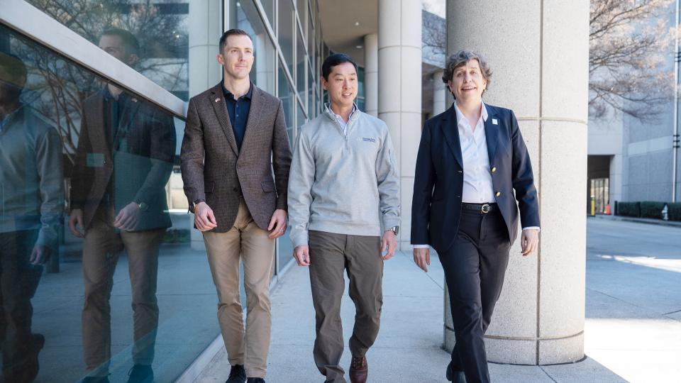 Three people walking together outside a modern building with large glass windows and concrete columns.