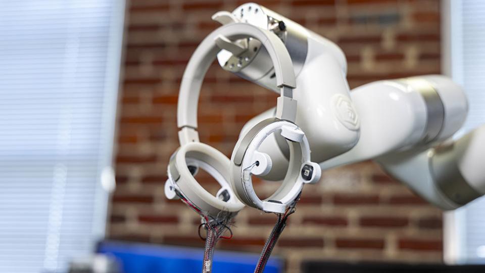 Robotic arm holding circular sensor devices with exposed wiring in a lab setting with a blurred brick wall background.
