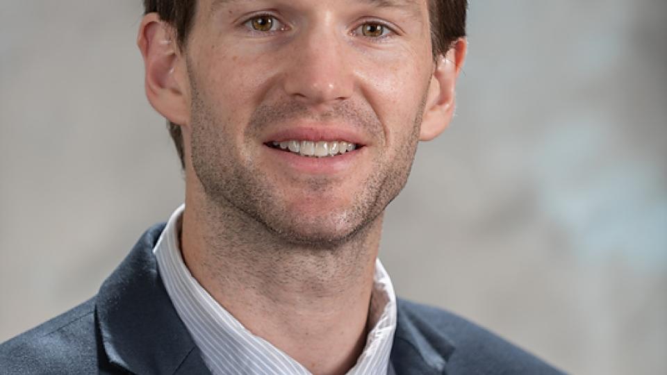 A man with brown hair and a short beard smiles for a portrait while wearing a dark blue suit and red tie.