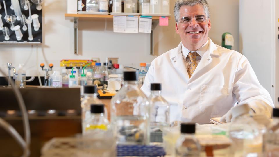 A man with silver hair wears a white lab coat, white shirt, and gold tie will sitting behind a lab bench with research equipment on top of it.