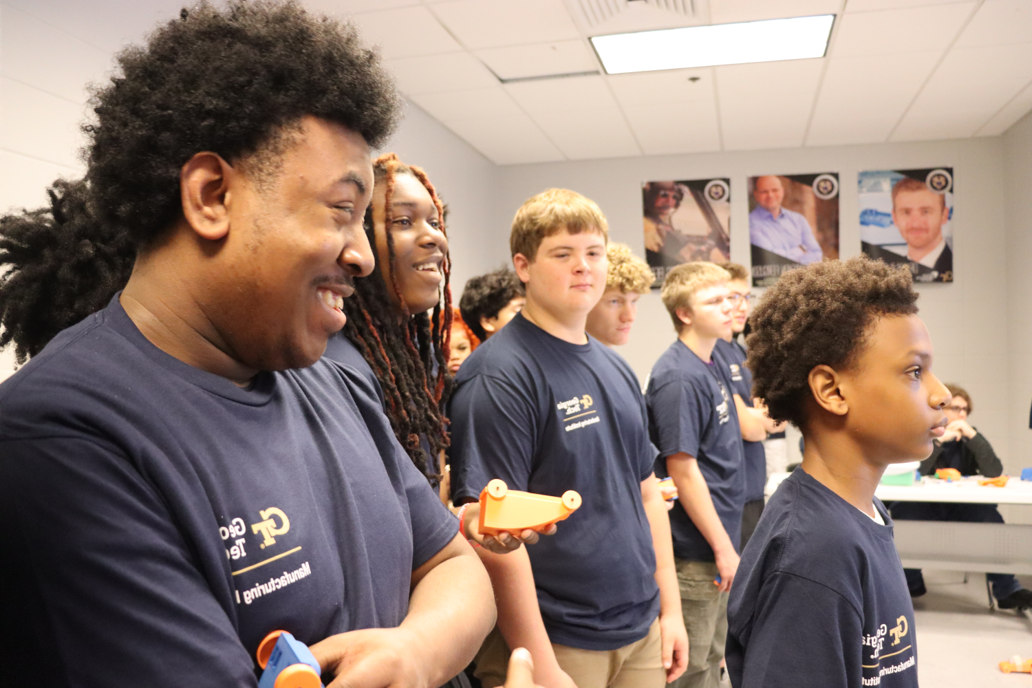 Group of students in navy shirts standing in a classroom, each holding small orange and blue 3D-printed objects.