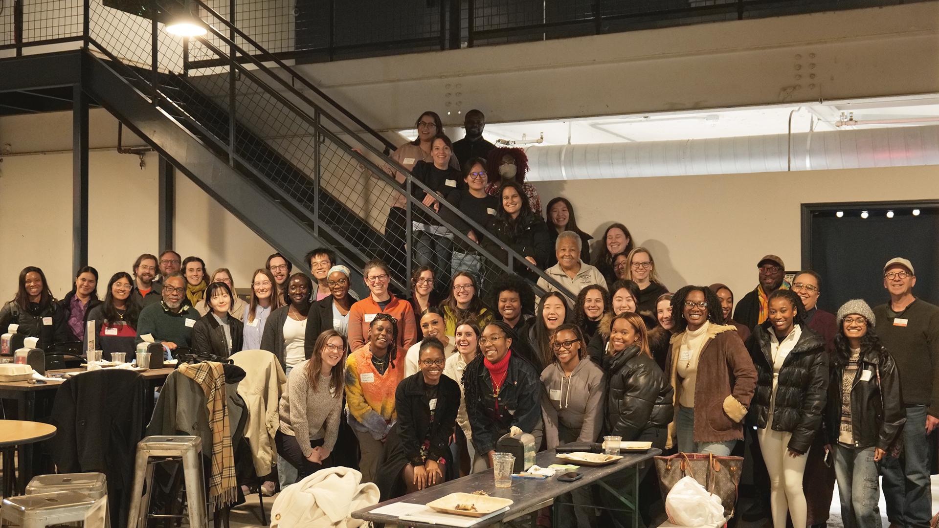 Large group of people standing and seated in a bright industrial-style indoor space, gathered on and around a metal staircase and long tables. The setting includes exposed beams, railings, overhead lighting, and tables with notebooks, cups, and coats visible in the foreground.