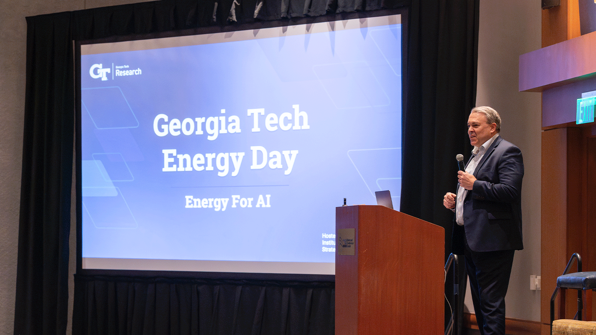 A man stands at a podium speaking in front of a large screen displaying “Georgia Tech Energy Day: Energy for AI.” The setting is a conference room with stage lighting and an audience out of frame.