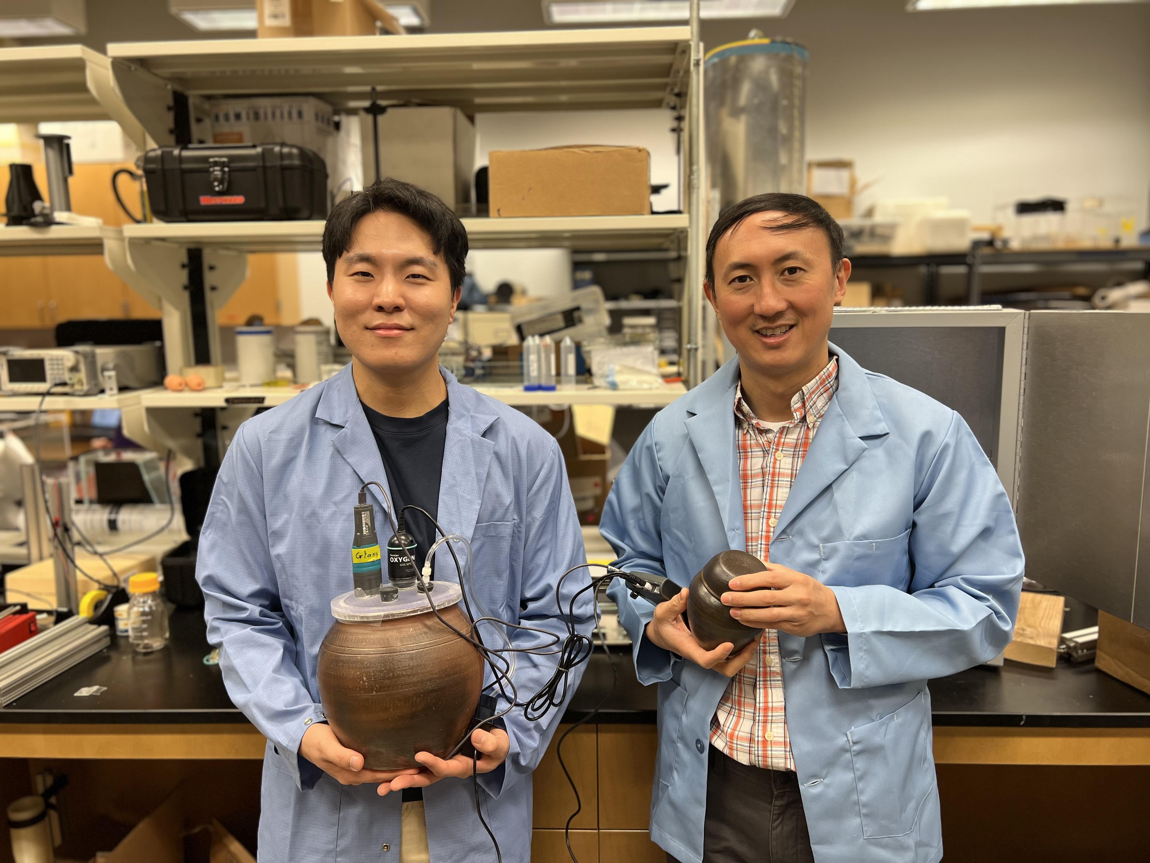 Two people in blue lab coats holding onggi - traditional Korean earthenware pots. 