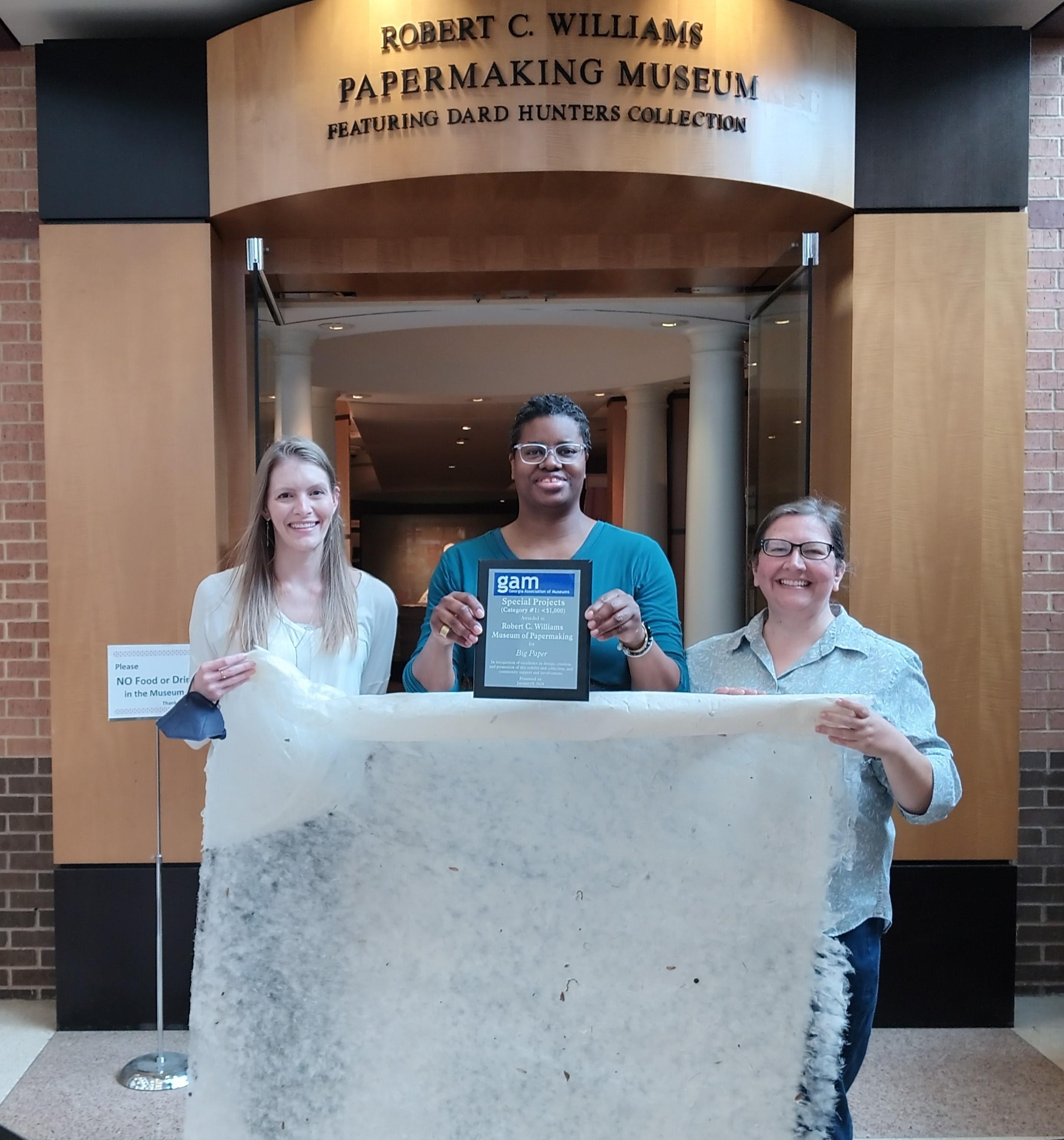 Three museum staff stand in front of the museum doors holding a very large sheet of paper and a plaque. 
