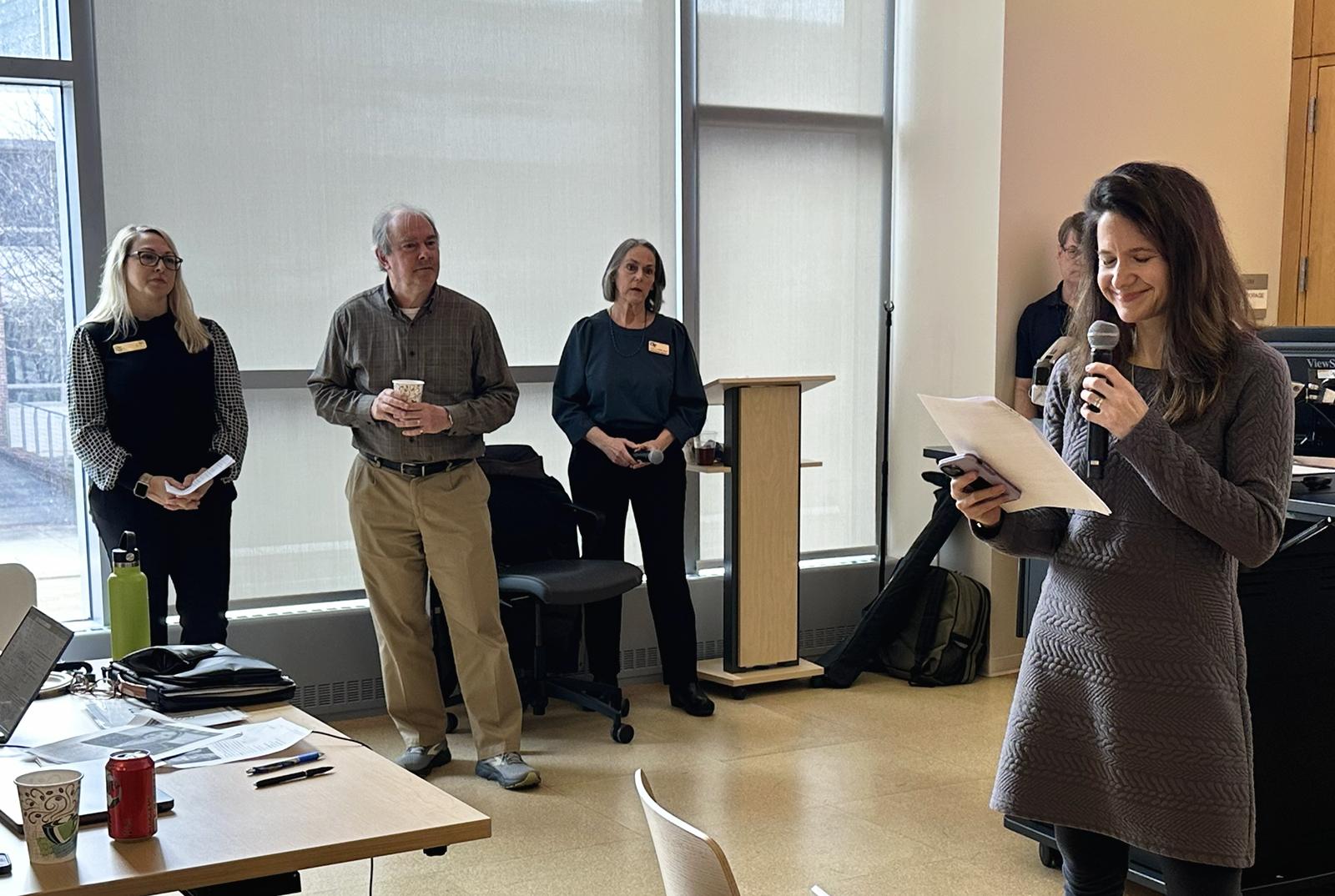 "A group of people stand near large windows in a bright indoor space, with one person holding a microphone and reading from a sheet of paper. Three individuals stand together near a lectern, some holding notes or drinks. In the foreground, a table holds laptops, papers, and a drink cup. The room has light-colored floors and tall window shades that diffuse the natural light."