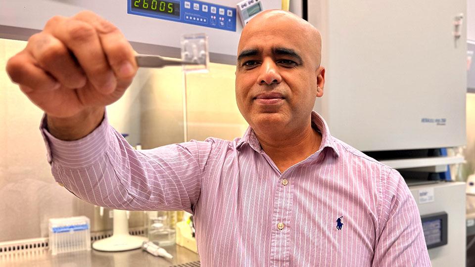 A researcher holds a plastic chip up to the camera while sitting in a lab setting in front of a fume hood.