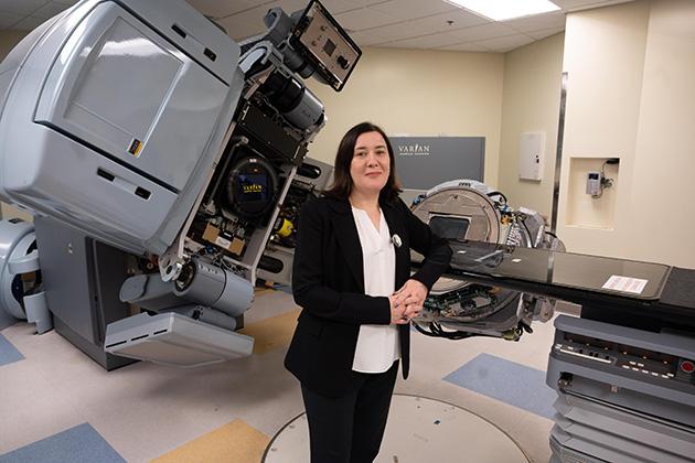 Person standing in a medical treatment room with advanced radiotherapy equipment, including a large Varian machine and treatment table.