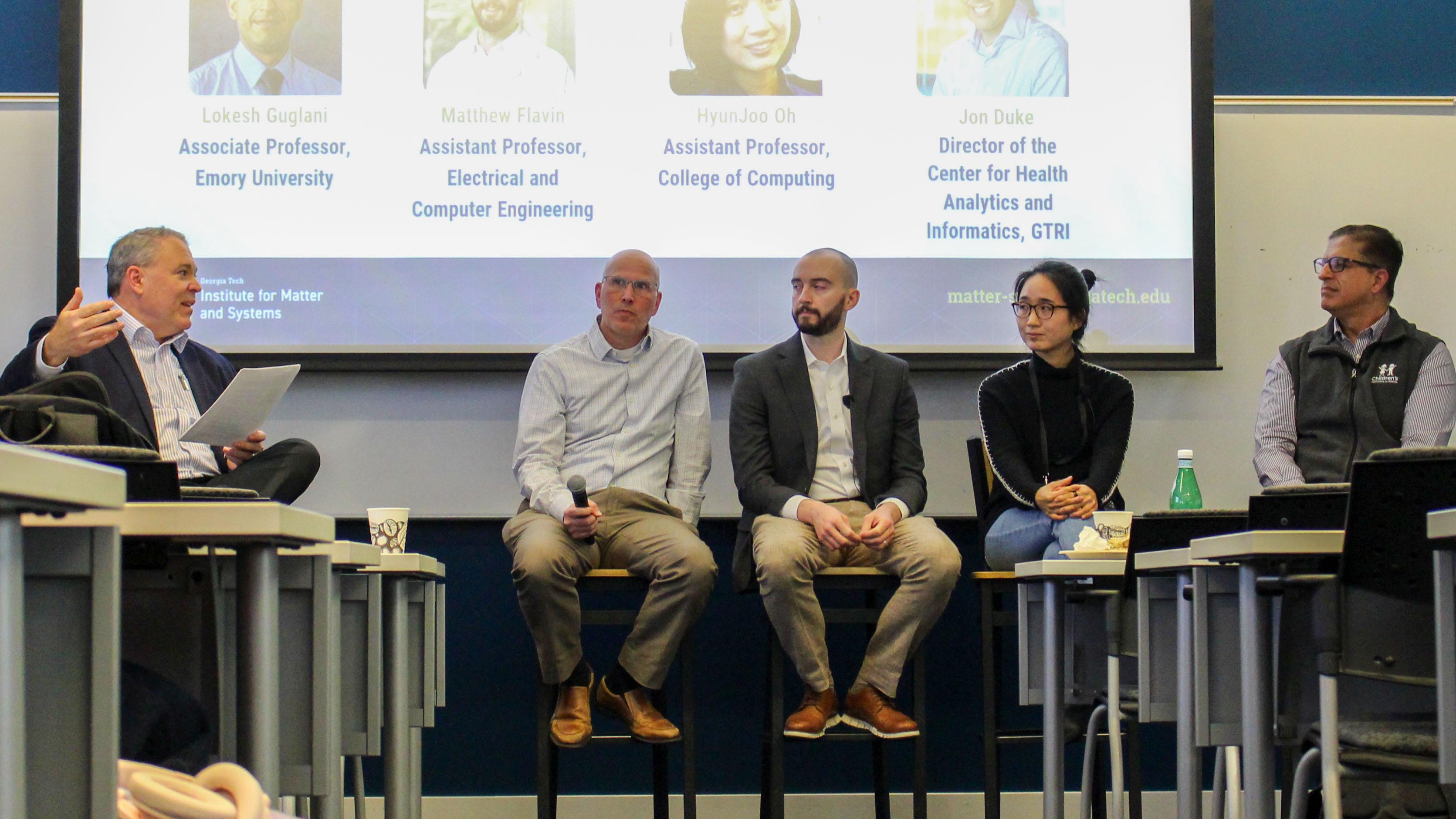 A panel of five speakers sits on tall stools at the front of a classroom, participating in a moderated discussion. The moderator on the left holds papers while addressing the group. A large presentation slide behind the panel displays names and academic titles. Audience members are partially visible in the foreground, and tables, chairs, and a water bottle are arranged throughout the room.