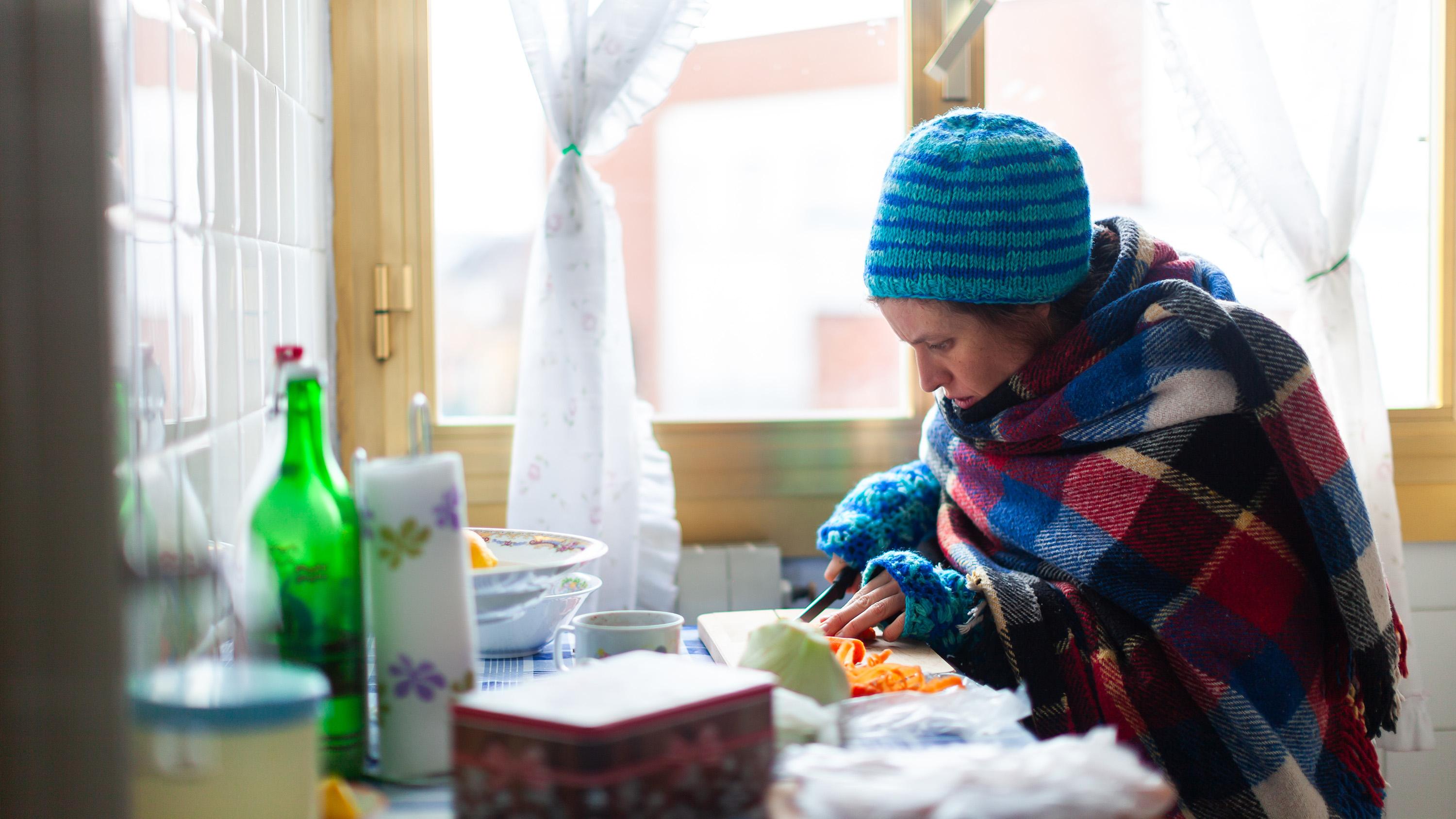 A woman wearing a hat and warm clothing prepares food in her kitchen.