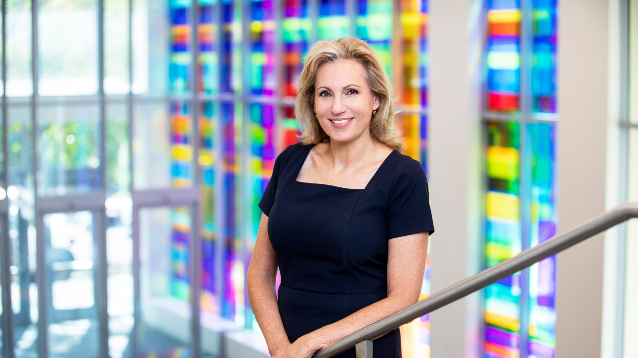 Laura Taylor, a blonde woman with a fair complexion, stands in a navy dress in front of a large window with a vibrant colored opaque coverings.