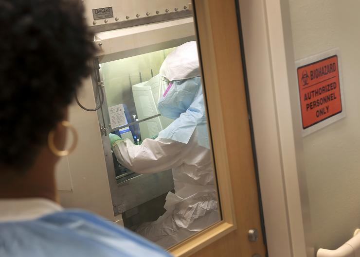 <p>Image shows a lab technician in Emory’s Serious Communicable Diseases Unit laboratory, with a team member watching from the outside for safety purposes. (Credit: Jack Kearse, Emory Health Sciences Photography)</p>