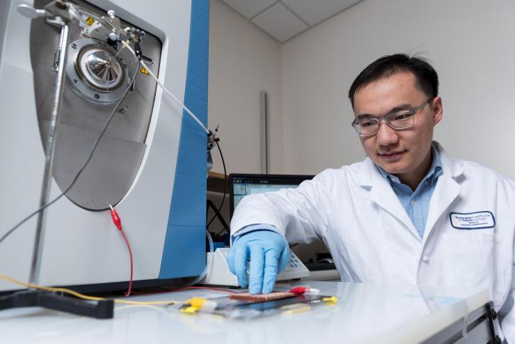 <p>Anyin Li, a postdoctoral fellow in the Georgia Tech School of Chemistry and Biochemistry, demonstrates the use of a sliding triboelectric nanogenerator to produce electrical charges for the mass spectrometer device shown next to him. (Credit: Rob Felt, Georgia Tech)</p>