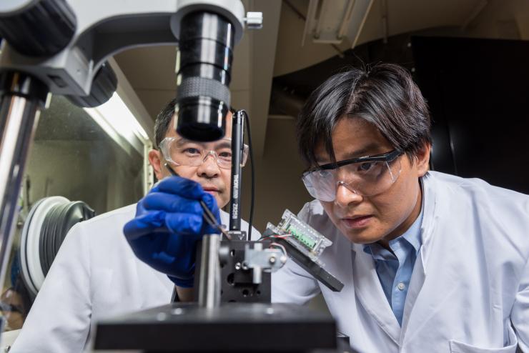 <p>(Left to right) Professor Ting Zhu and Assistant Professor Shuman Xia, both from Georgia Tech’s Woodruff School of Mechanical Engineering, show how a thin film electrode made of amorphous silicon was tested in a custom environmental indenter. To provide proper environmental control, samples containing lithiated silicon were tested with the device inside the glovebox shown in the background. (Credit: Rob Felt, Georgia Tech)</p>