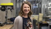 Woman in a workshop environment with industrial equipment and tools in the background, wearing a floral-patterned blouse and light knit cardigan, representing a modern manufacturing or maker space setting.