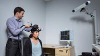 Researcher adjusting a device on another person’s head in a lab, with a computer displaying brain imaging data and a mounted camera in the background.