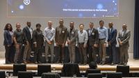 Group of professionals standing on a stage in front of a large presentation screen displaying agenda and seed grant booklet information at a conference.