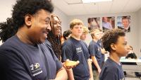 Group of students in navy shirts standing in a classroom, each holding small orange and blue 3D-printed objects.
