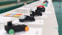 A row of small, student‑designed model race cars displayed on a table, each placed on top of design sheets in a gym setting.