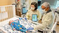 Two adults wearing protective gowns and gloves stand beside a hospital crib, using a tablet device while examining an infant lying on the mattress as medical equipment and monitors surround the crib.