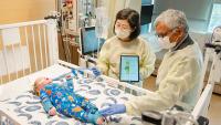 Two adults wearing protective gowns and gloves stand beside a hospital crib, using a tablet device while examining an infant lying on the mattress as medical equipment and monitors surround the crib.