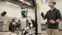 Person wearing a virtual reality headset controlling a humanoid robot equipped with tools in a laboratory setting.