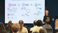 A presenter stands at the front of a lecture room speaking to a seated audience while a projected slide titled “Synthetic Biology: Engineered Gene Circuits” illustrates the design–build–test cycle with diagrams and icons explaining gene circuit construction and testing.
