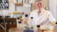 A man with silver hair wears a white lab coat, white shirt, and gold tie will sitting behind a lab bench with research equipment on top of it.