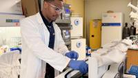 A man in a lab coat wearing safety goggles and gloves puts samples into a machine in a scientific lab