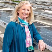 A woman with blonde hair and a blue sweater stands among solar panels. 