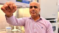 A researcher holds a plastic chip up to the camera while sitting in a lab setting in front of a fume hood.