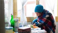 A woman wearing a hat and warm clothing prepares food in her kitchen.