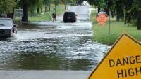 A car drives through a flooded section of a neighborhood street.