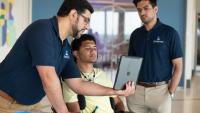 Three people review a tablet together in a lab setting, with one seated individual using a powered wheelchair and assistive device.
