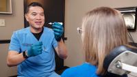 Person in blue medical scrubs demonstrates a small device to a seated patient in a clinical exam room with medical equipment visible in the background.