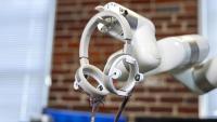 Robotic arm holding circular sensor devices with exposed wiring in a lab setting with a blurred brick wall background.