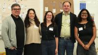 Five people stand side by side in a meeting or workshop space with large planning boards displayed behind them. The boards are covered with flowcharts, diagrams, handwritten notes, and sticky notes outlining process maps and project planning information. The group is positioned in front of the displays, with name tags visible.